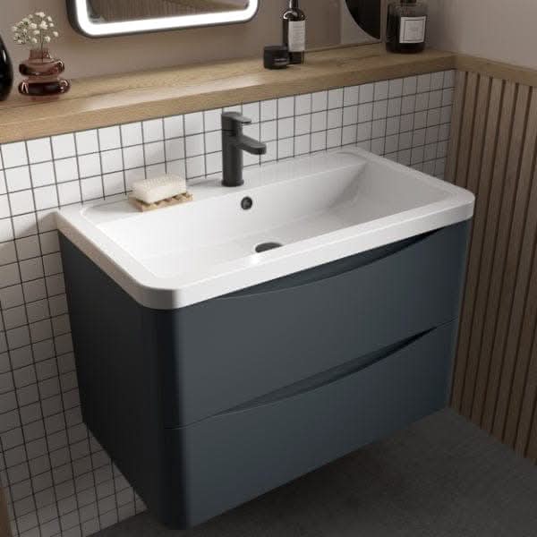 The Nuie Lunar 2 Drawer Wall Hung Vanity Unit with Ceramic Basin (600mm), in dark gray, features two drawers and a modern rectangular basin by Nuie, complemented by a wall-mounted faucet, soap dish, and toiletries on a shelf above white tiles.