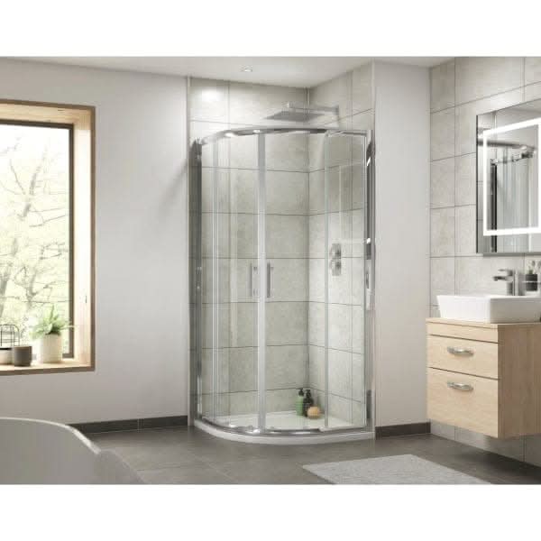 A modern bathroom featuring a Nuie Pacific Quadrant Shower Enclosure in chrome, tiled walls, a wooden vanity with white sink, wall mirror, and large window. Toiletries are visible inside the Nuie shower enclosure.