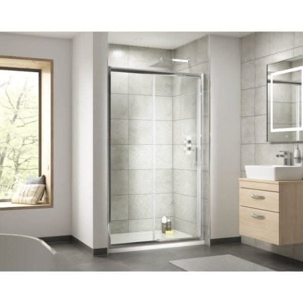 Modern bathroom featuring a Nuie Pacific Sliding Shower Door in chrome, gray tiled walls, wooden vanity with white sink, large mirror, window with a view of trees, and two bottles on the shower floor.