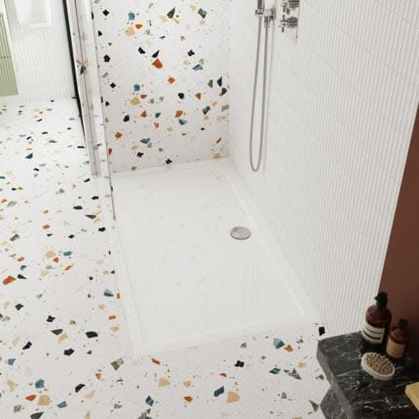 A modern bathroom with a walk-in shower featuring the Nuie Pearlstone Rectangular Shower Tray in white, terrazzo floor and wall tiles with colorful chips, white tiled wall, and toiletries on a dark countertop.