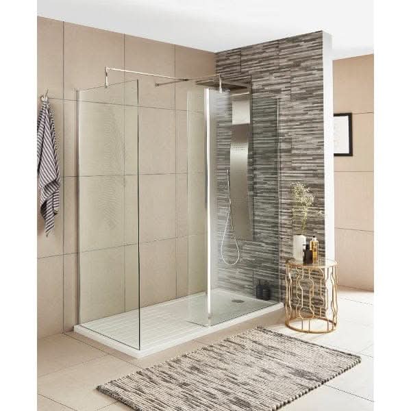 Modern bathroom with a glass-enclosed walk-in shower featuring the Nuie Wetroom Return Screen by Nuie and a rainfall showerhead. A striped rug, gold side table with vase, beige and gray tiles, and a sleek vertical mirror complete the space.