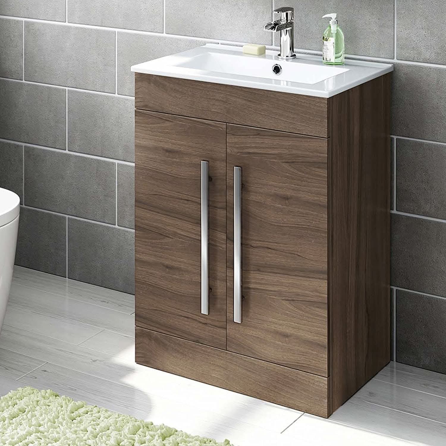 The iBathUK Avon 2 Door Floorstanding Vanity Unit With Basin (615mm x 815mm, 1 Tap Hole) features a white basin atop a stylish wood cabinet with two long metal handles. Chrome tap and soap dispenser included, set against gray tiles with a green rug nearby.
