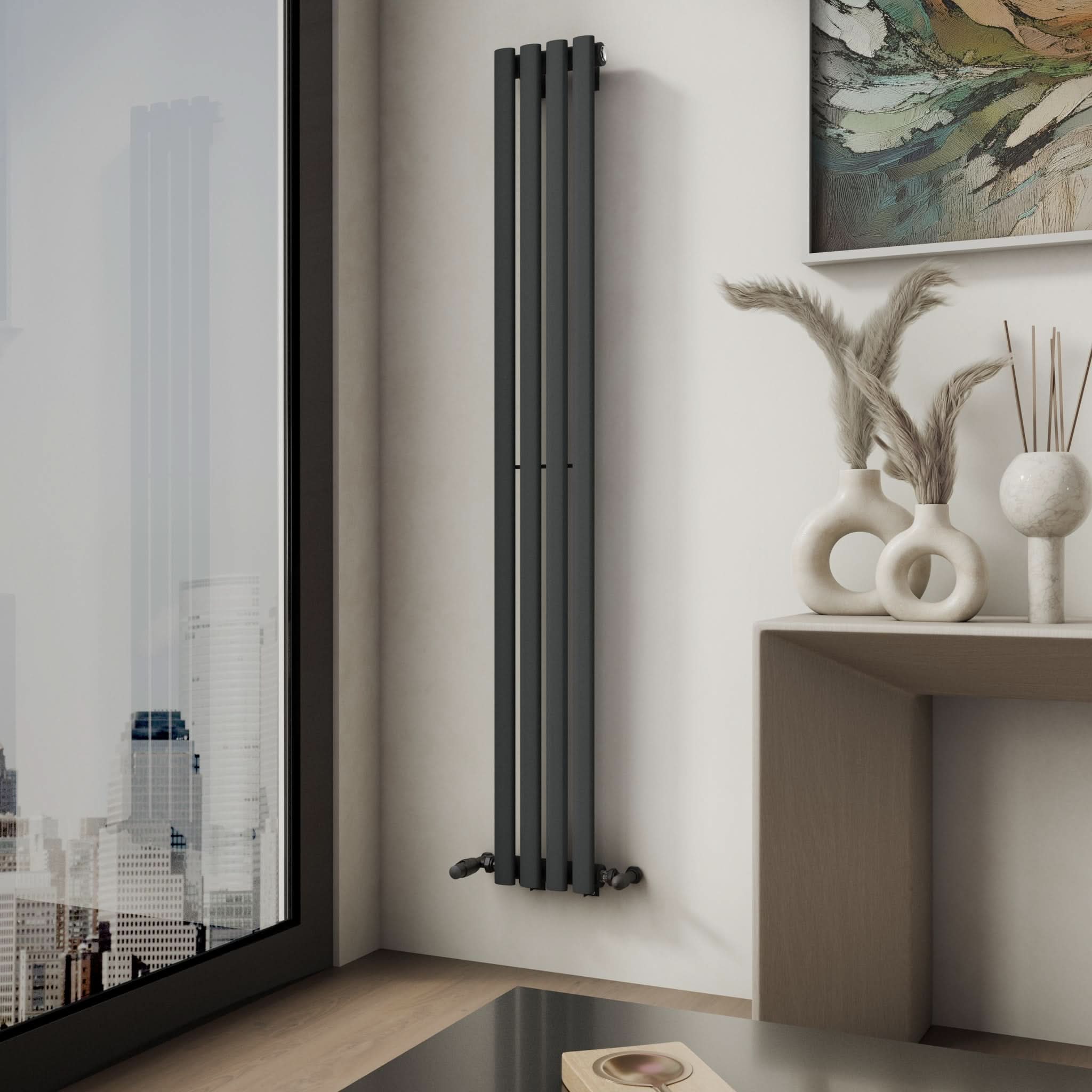 A modern room corner features the iBathUK Princia Vertical Single Oval Panel Radiator on a white wall beside a city-view window. A table displays vases with dried grasses, and an abstract painting above adds a stylish accent.