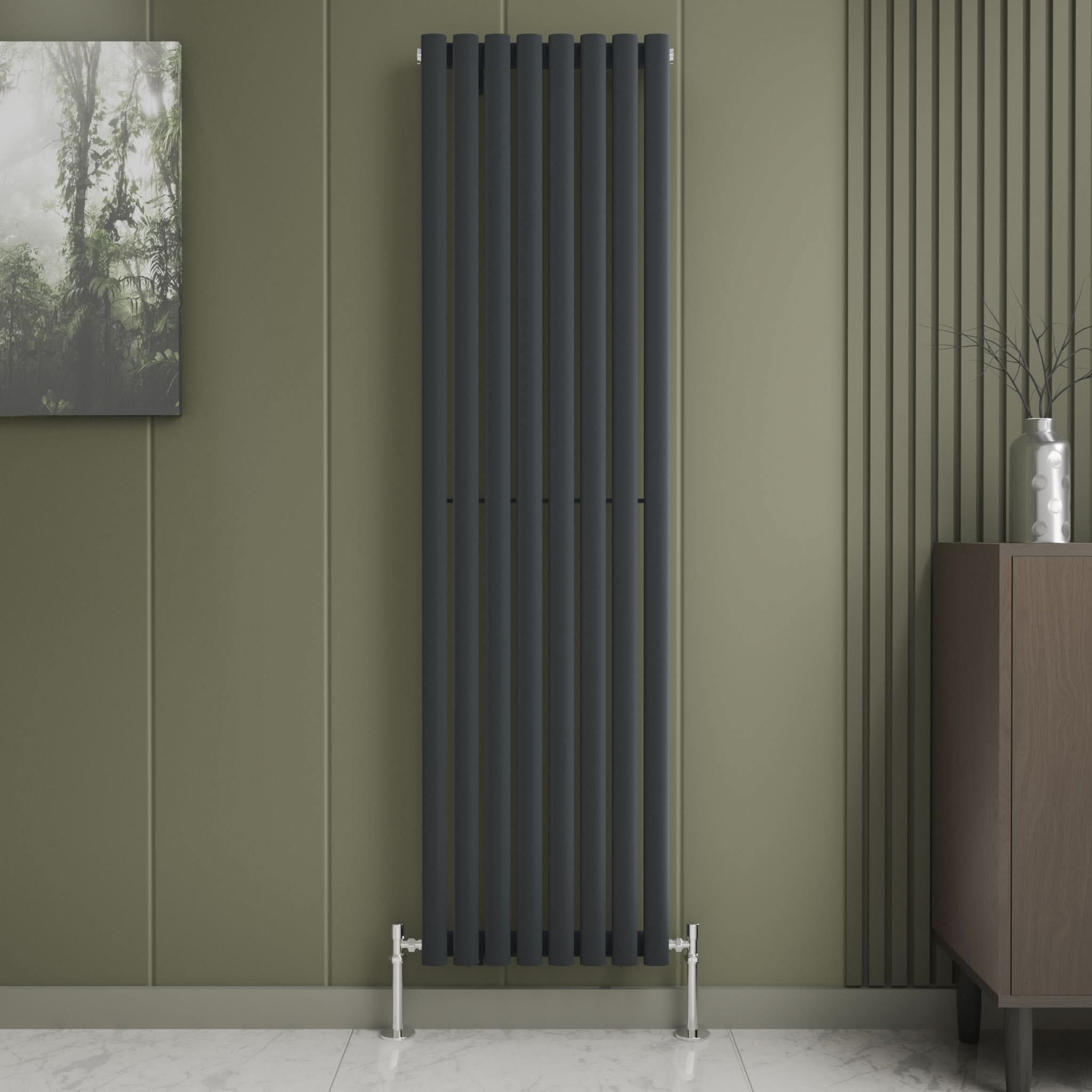 The iBathUK Princia Vertical Single Oval Panel Radiator is mounted on an olive-green wall beside decorative panels. A small cabinet with a vase stands to the right, with a framed nature print to the left above white tile flooring.