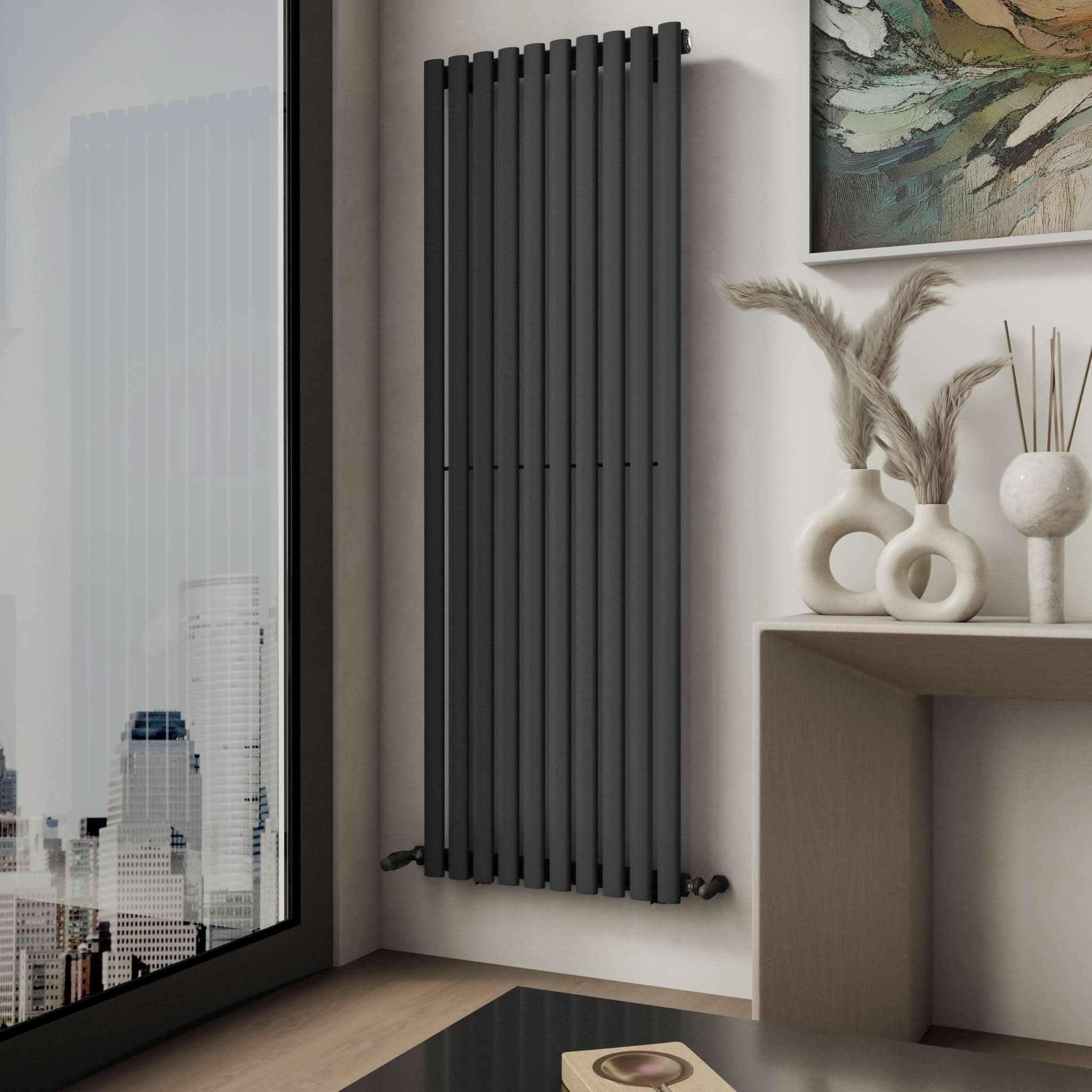 A Princia Vertical Single Oval Panel Radiator by iBathUK in black is wall-mounted beside a large window with city views, while a light console table topped with decorative vases and dried grass enhances the stylish setting.
