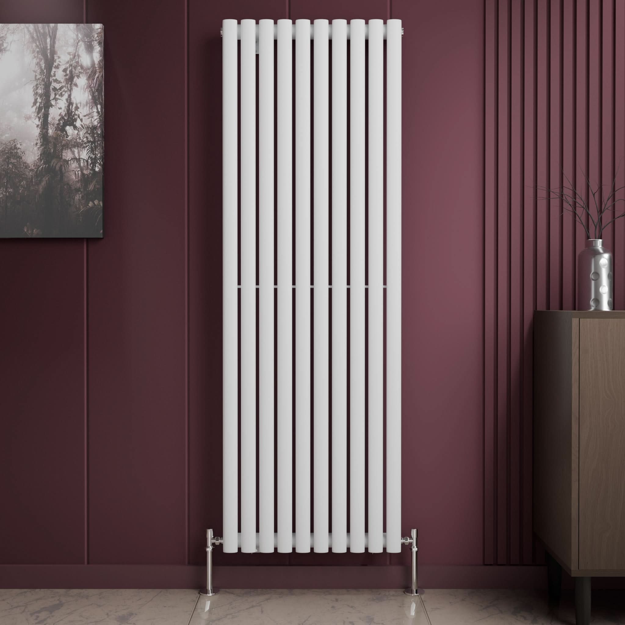 The iBathUK Princia Vertical Single Oval Panel Radiator in white is mounted on a dark maroon wall. To its right stands a brown cabinet with a silver vase; to the left, a framed picture. The floor features light-colored tiles.