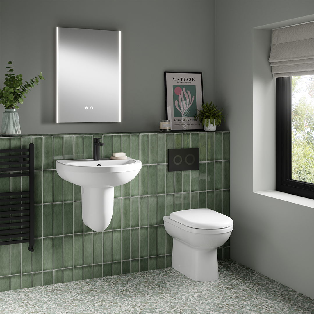 Modern bathroom featuring a green tiled wall, white wall-mounted sink, and the Nuie Ivo Back to Wall Toilet by Nuie. A black towel heater, illuminated mirror, framed art, potted plants, and a window with Roman shade complete the stylish look.