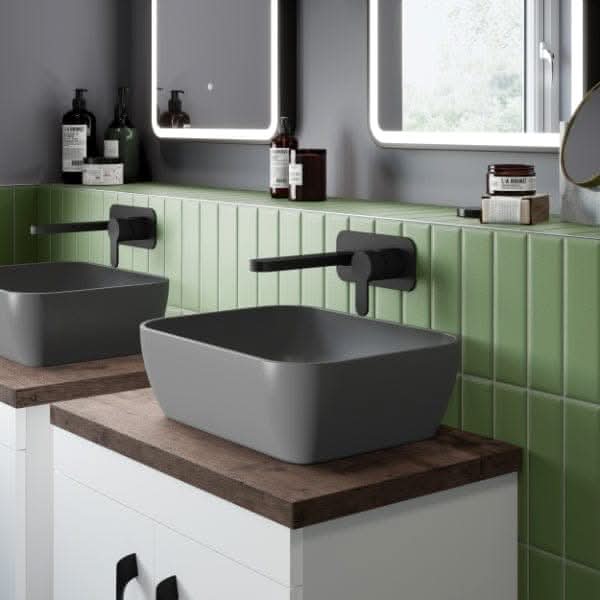 Contemporary bathroom featuring two Nuie 455mm Sit-On Countertop Basins by Nuie on wooden countertops, green tiled backsplash, wall-mounted black faucets, and a large LED-lit mirror. Bottles and toiletries are neatly arranged near the basins.